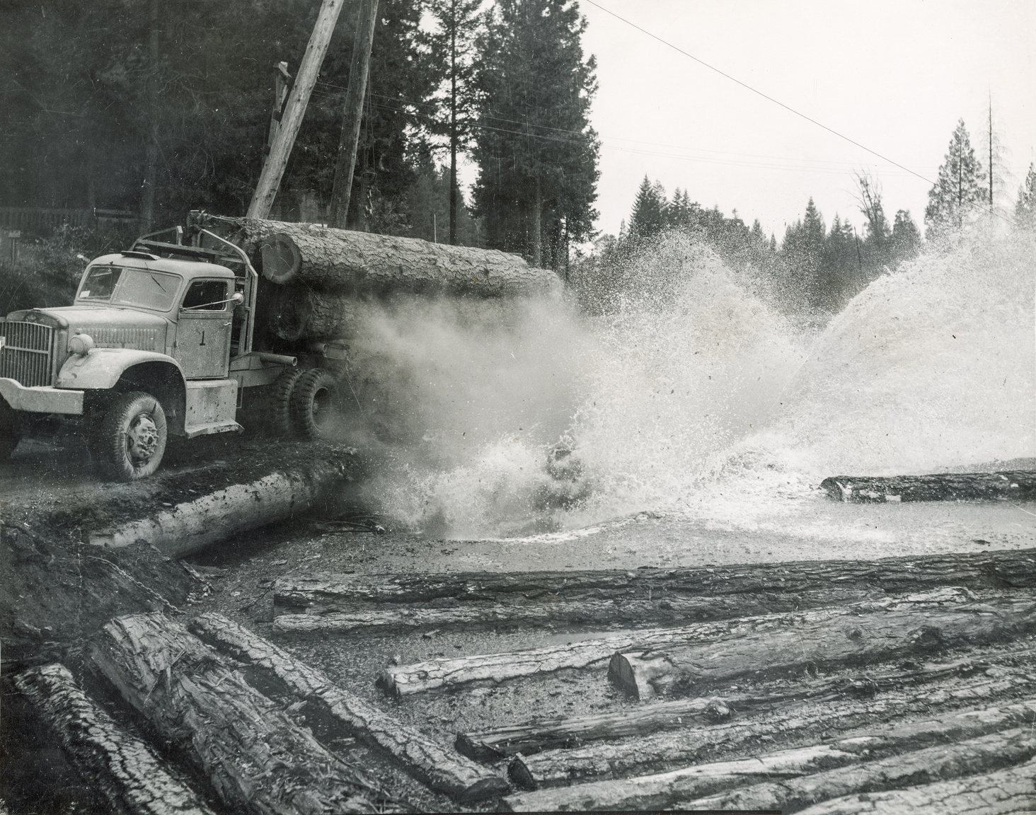 Log truck at a landing with water spraying across the deck to control dust, circa 1950