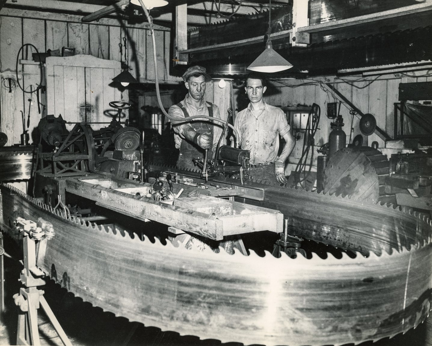Two men filing a large circular head saw inside a mill filing room, circa 1950